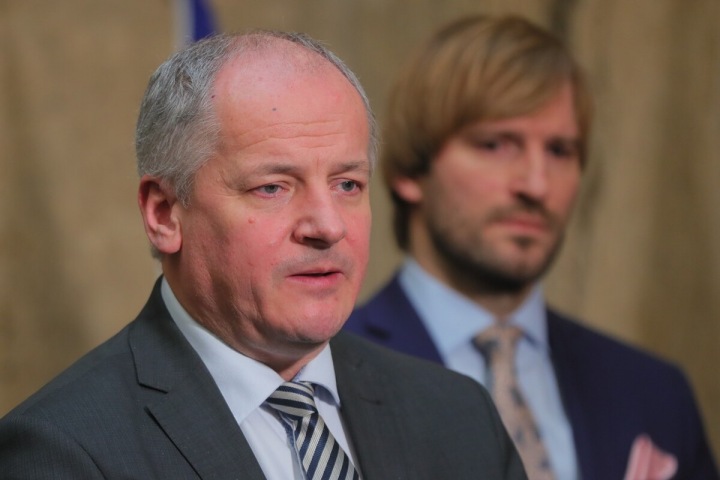 Roman Prymula stojí v šedém obleku, za ním Adam Vojtěch